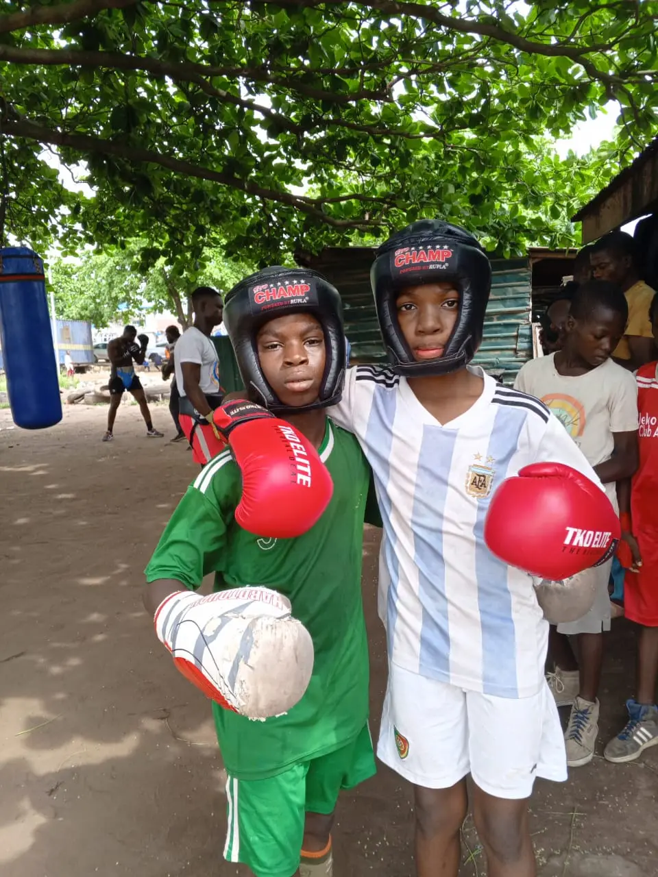 Two young boxers in headgear and red gloves, grinning under a tree at training